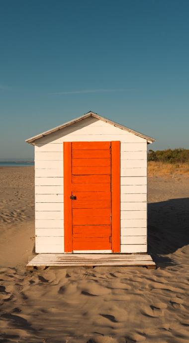 Cabina bianca con porta arancione su una spiaggia deserta al tramonto.