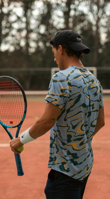 Giocatore di tennis con racchetta e pallina su campo all'aperto, indossa cappello e maglietta.