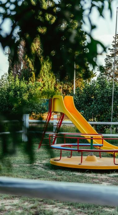 Parco giochi con scivolo giallo e giostra, circondato da alberi e verde.