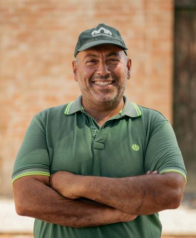 Uomo sorridente con cappello e polo verde, braccia incrociate, sfondo di mattoni.