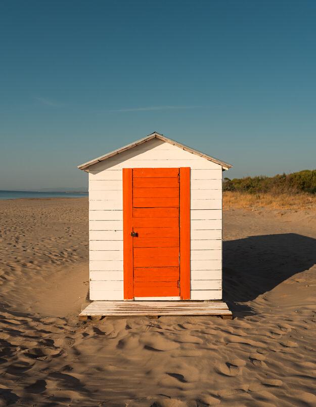 Cabina bianca con porta arancione su una spiaggia deserta al tramonto.