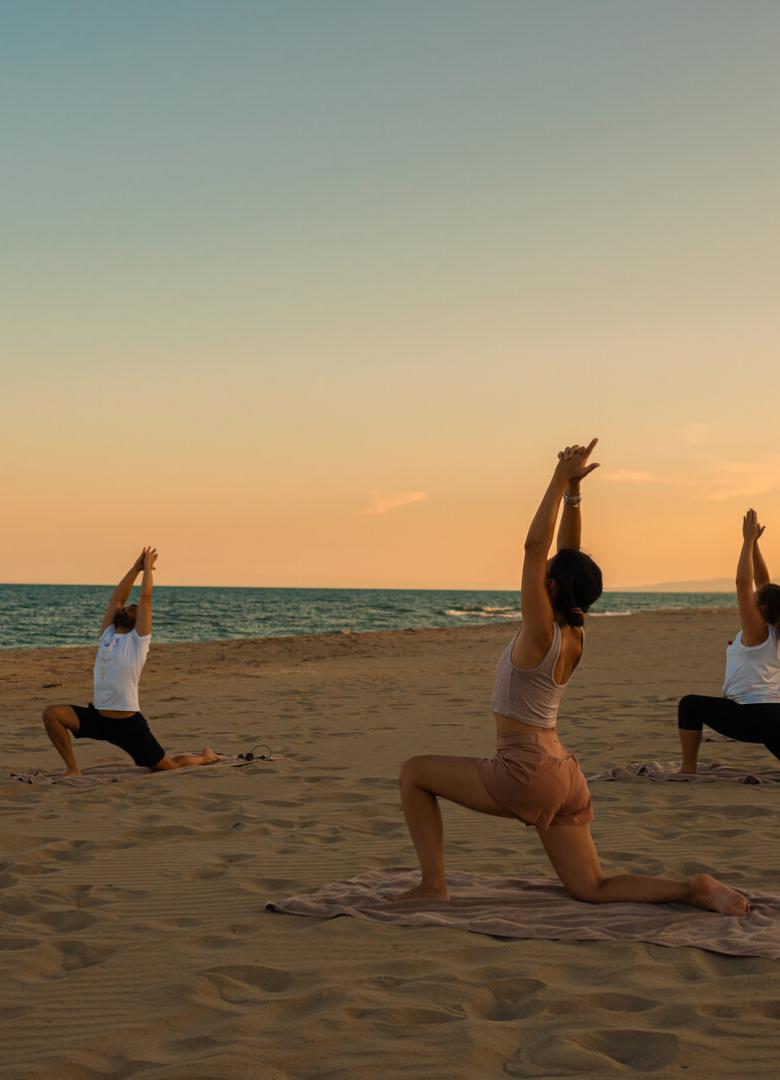 Persone praticano yoga al tramonto su una spiaggia, in pose di meditazione.