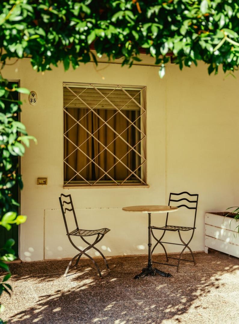 Piccolo patio con tavolo e sedie in metallo, ombreggiato da piante verdi.