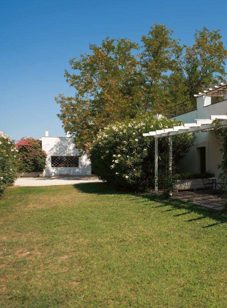 Casa bianca con veranda, giardino e piante rampicanti sotto un cielo azzurro.